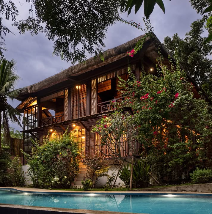 The Red Hen Homestead at dusk — two-storey Filipino heritage farmhouse with cogon thatched roof, lap pool, and bougainvillea, under a moody purple-blue evening sky at Mt. Banoy, Batangas City